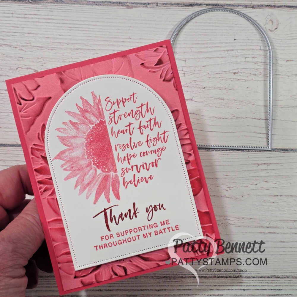 A hand holds a pink, handmade card with an embossed floral background. The card features a large pink sunflower, uplifting script text, and a thank you message for support during a battle. A metal die-cut arch shape lies on the wooden surface beside the card. To read more, visit Patty Stamps (pattystamps.com)