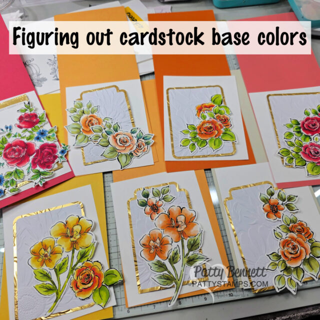 A workspace with handmade floral cards on orange, yellow, and coral cardstock. Each card features colorful stamped and colored flowers with gold foil borders. Text at the top reads "Figuring out cardstock base colors." Craft tools are visible in the background. To read more, visit Patty Stamps (pattystamps.com)
