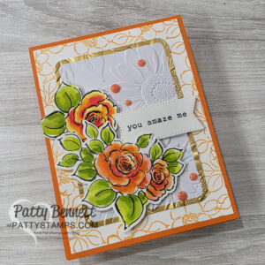 A handmade greeting card with orange border, floral embossed white panel, and gold accents. Watercolored orange roses with green leaves, pink enamel dots, and a white stitched label saying “you amaze me.” Card is signed “Patty Bennett, PattyStamps.com.”. To read more, visit Patty Stamps (pattystamps.com)