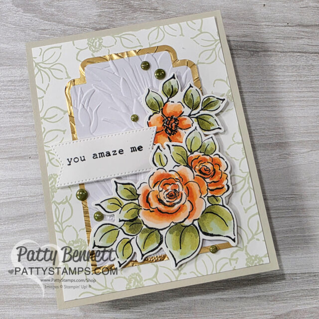 A handmade card features hand-colored orange and yellow roses with green leaves, accented with gold foil and green gems. Embossed white paper and a “you amaze me” sentiment are layered on a pale floral background. The card rests on a light wood surface. To read more, visit Patty Stamps (pattystamps.com)