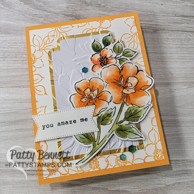 A handmade card features orange watercolor flowers with green leaves layered on embossed white and gold-patterned papers. A stitched banner reads “you amaze me.” Blue gem embellishments and the “Patty Bennett PattysStamps.com” watermark are visible. To read more, visit Patty Stamps (pattystamps.com)