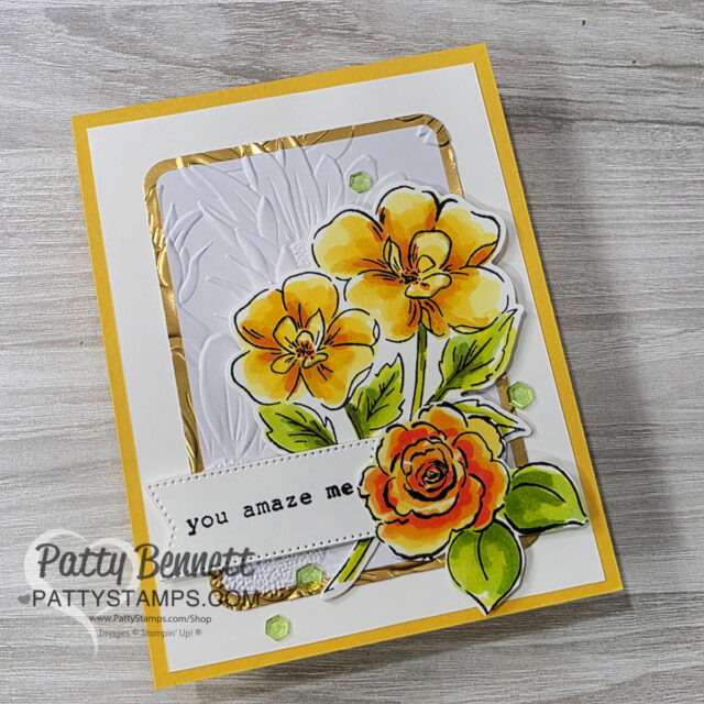 A handmade greeting card with yellow and orange flowers, green leaves, and embossed white background bordered in gold. A white tag with “you amaze me” in black type and green sequins decorate the card. The creator’s watermark, “Patty Bennett,” is at the bottom left. To read more, visit Patty Stamps (pattystamps.com)