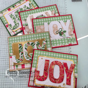 Six handmade holiday cards are arranged on a cutting mat. Each card features the word "JOY" in large, decorative letters with festive patterns, including florals, greenery, and gold accents. Backgrounds are layered with green, red, and plaid designs. To read more, visit Patty Stamps (pattystamps.com)