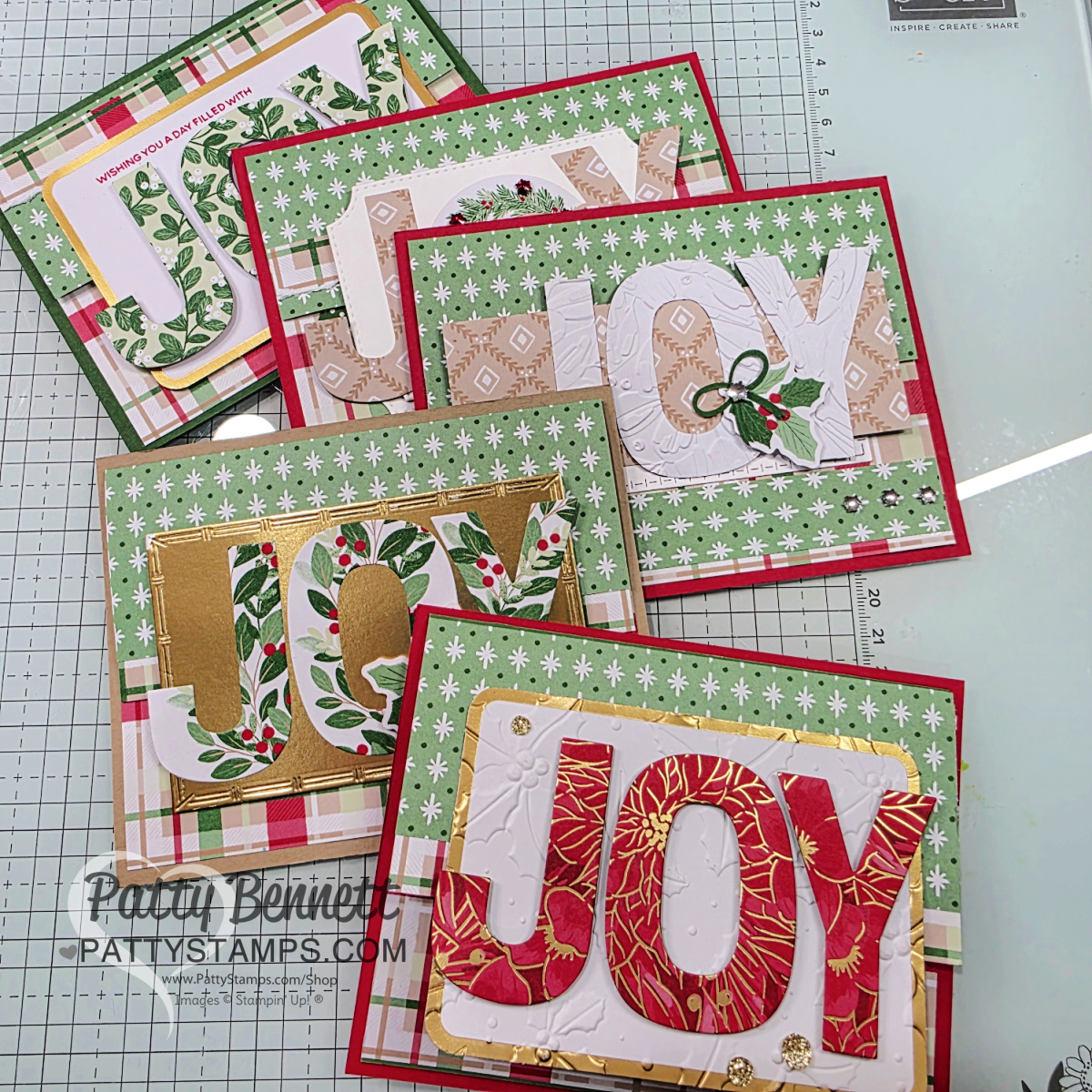 Six handmade holiday cards are arranged on a cutting mat. Each card features the word "JOY" in large, decorative letters with festive patterns, including florals, greenery, and gold accents. Backgrounds are layered with green, red, and plaid designs. To read more, visit Patty Stamps (pattystamps.com)