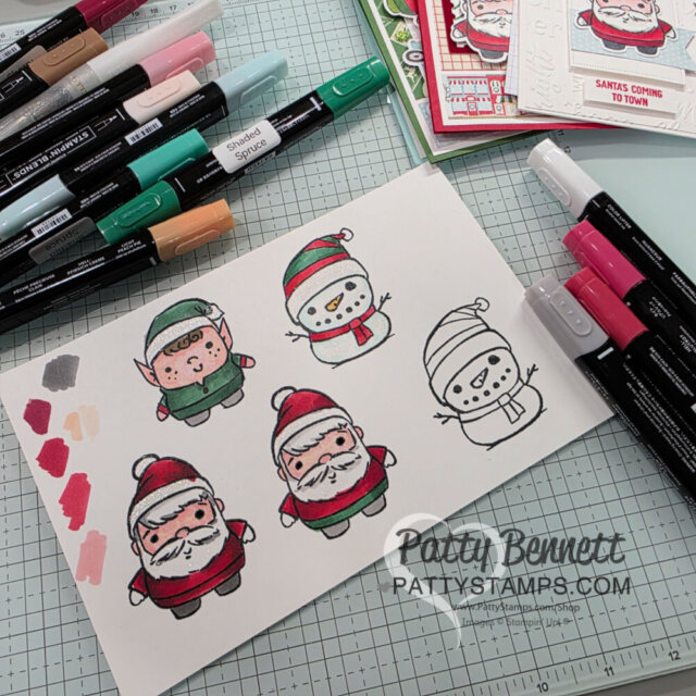 A sheet of paper shows colored drawings of two elves, two snowmen, and two Santas. Various markers are scattered around, with some color swatches on the paper. Handmade Christmas cards are visible in the background, and a visible watermark reads "PattyStamps.com. To read more, visit Patty Stamps (pattystamps.com)