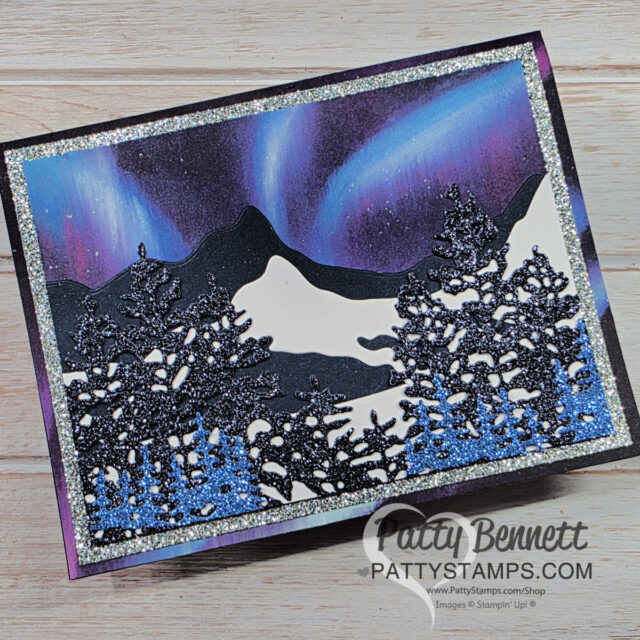A handmade greeting card features glittery black die-cut trees in the foreground, with snowy hills, distant black mountains, and a vibrant aurora borealis in purple, blue, and green tones in the sky. The card has a sparkling blue border and “PattyStamps.com” watermark. To read more, visit Patty Stamps (pattystamps.com)