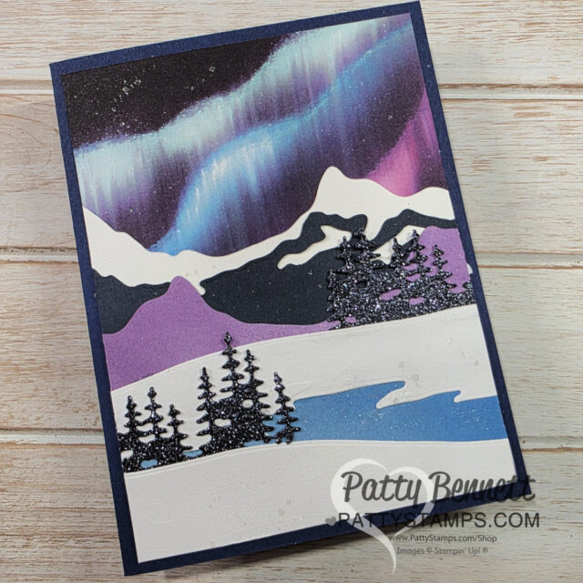 A handmade greeting card features snowy hills, black glitter pine trees, and a vibrant aurora borealis sky in purple, blue, and white. The card has layered paper cutouts for the landscape and is displayed on a light wood surface. “Patty Bennett” watermark is visible. To read more, visit Patty Stamps (pattystamps.com)
