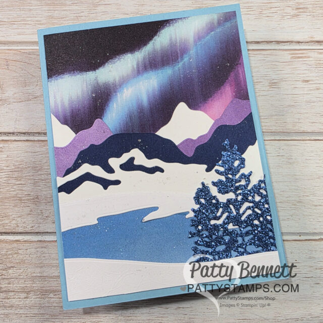 A handmade greeting card featuring a snowy mountain landscape with sparkling blue trees in the foreground and a colorful aurora borealis in purple, blue, and white hues in the night sky; “Patty Bennett pattystamps.com” is printed at the bottom right. To read more, visit Patty Stamps (pattystamps.com)