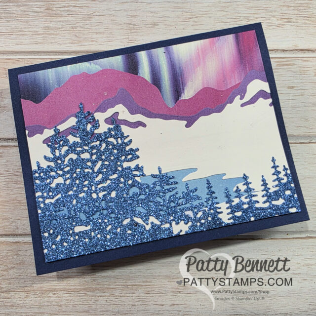A handmade card featuring glittery blue pine trees in the foreground, pink and purple mountains in the background, and a sky with aurora borealis-like streaks. The card is displayed on a wooden surface with a watermark reading "Patty Stamps. To read more, visit Patty Stamps (pattystamps.com)