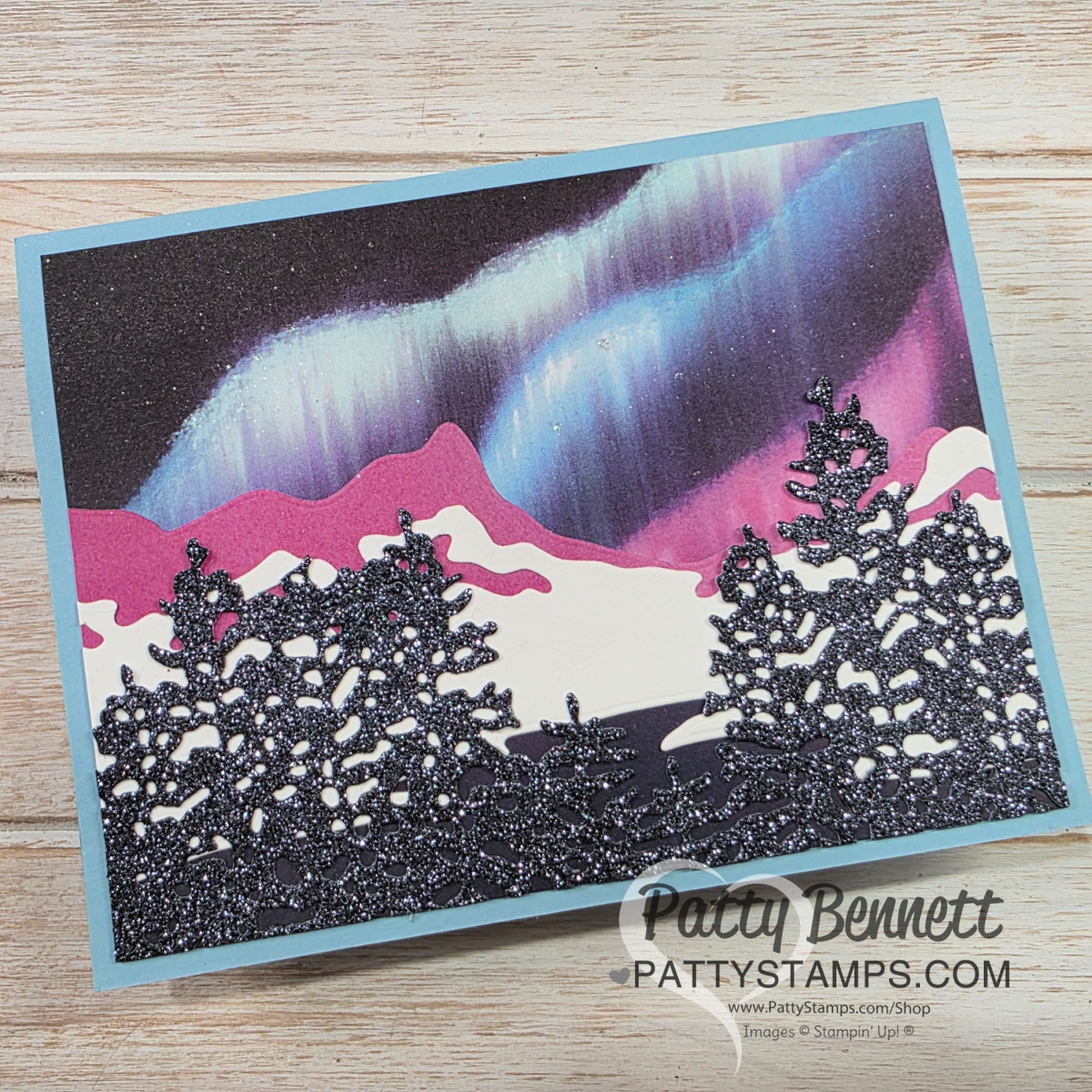 A handmade greeting card featuring a wintry night scene with silhouetted, glittery black pine trees in the foreground, snow-covered hills, and a vibrant aurora borealis in shades of blue, purple, and green across a dark sky. To read more, visit Patty Stamps (pattystamps.com)