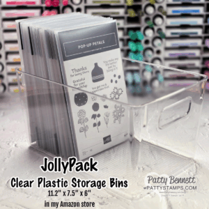 A clear plastic storage bin holds several upright crafting stamp sets labeled "Pop-Up Petals." The bin has a handle cutout and sits on a marble surface. Text on the image advertises "JollyPack Clear Plastic Storage Bins" with dimensions and Amazon store info. To read more, visit Patty Stamps (pattystamps.com)