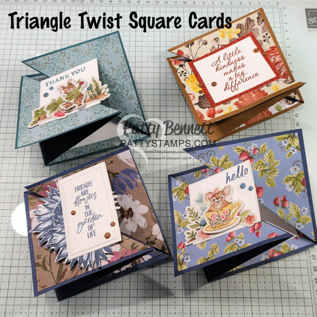 Four handmade Triangle Twist Square Cards with floral designs are arranged on a grid mat. Each card features decorative papers, stamped greetings, and small embellishments. The text reads "Triangle Twist Square Cards." The cards are colorful and layered. To read more, visit Patty Stamps (pattystamps.com)