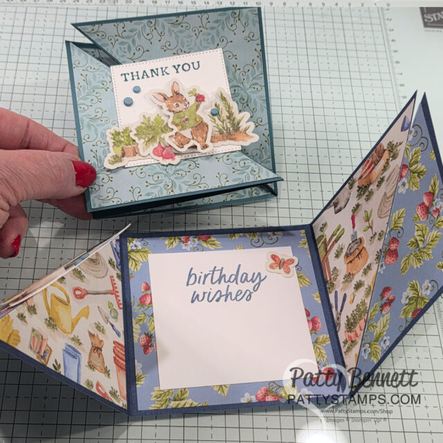 Two handmade pop-up cards are shown on a craft grid. One card has a “THANK YOU” message with a bunny, flowers, and garden scene on blue floral paper. The other reveals a “birthday wishes” note, surrounded by pastel garden-themed paper. A hand holds one card. To read more, visit Patty Stamps (pattystamps.com)