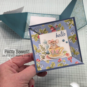 A hand holds a square handmade card with floral patterns, a cute mouse in a teacup, and the word "hello." The background features crafting supplies, including a partially open blue envelope. The card promotes pattystamps.com. To read more, visit Patty Stamps (pattystamps.com)