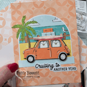 A handmade card features a cartoon orange car with two penguins inside, surfboards on the roof, a palm tree, beach, and a seagull in the background. The text reads "Cruising to Another Year." A hand holds the card on a patterned surface. To read more, visit Patty Stamps (pattystamps.com)