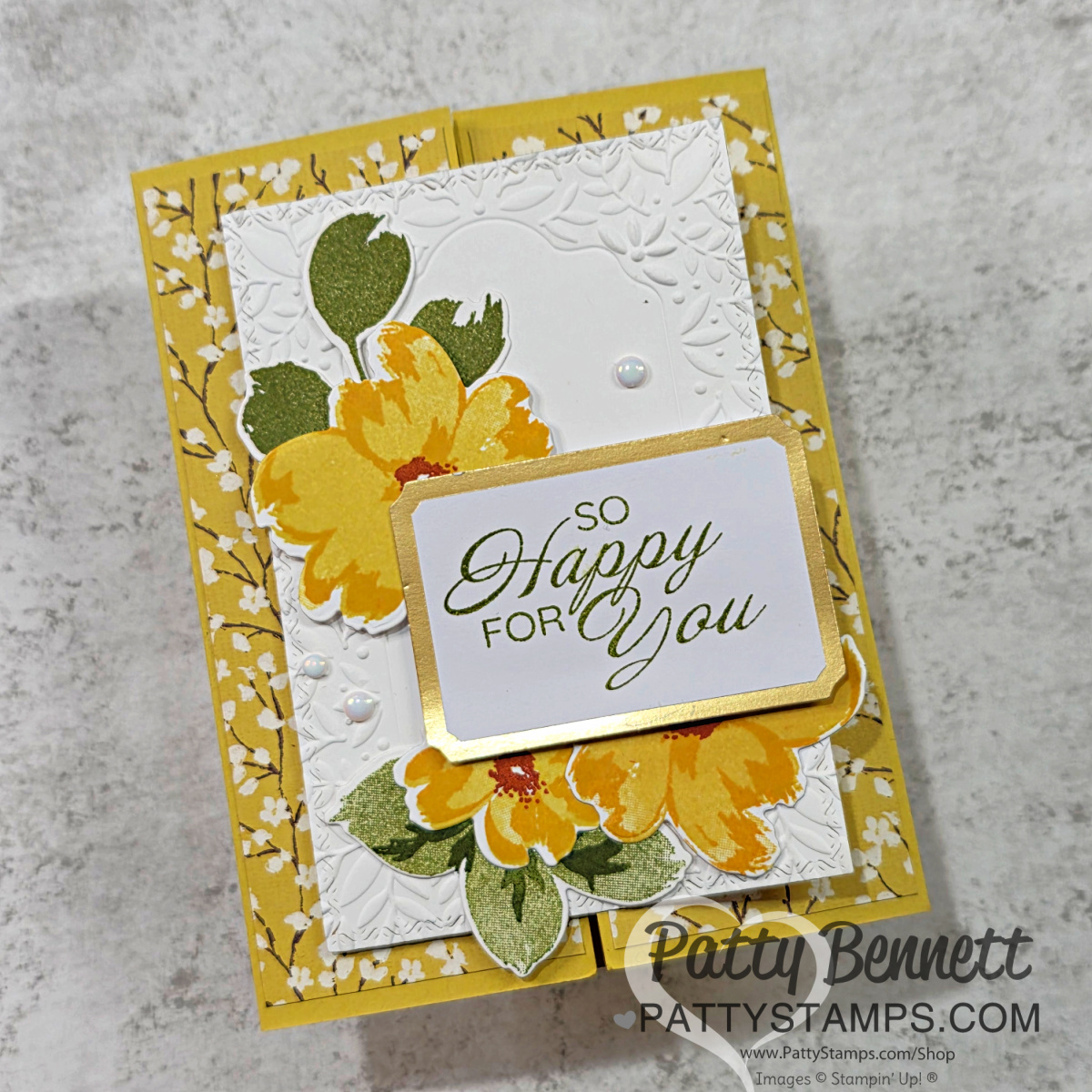 A handmade floral card with yellow and green flowers on an embossed white panel, bordered by gold. The card base is yellow with a white floral pattern. The sentiment reads “SO HAPPY FOR YOU” in green script. Pearls and leaves accent the design. To read more, visit Patty Stamps (pattystamps.com)