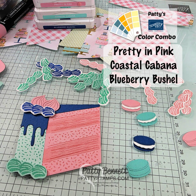 A craft workspace featuring die-cut paper treats, including a pink cake slice with blue and green icing and pink, teal, and blue macarons. Stamped and die-cut supplies, ink pads, and patterned papers are scattered around. Text highlights a pink, teal, and blue color combo. To read more, visit Patty Stamps (pattystamps.com)