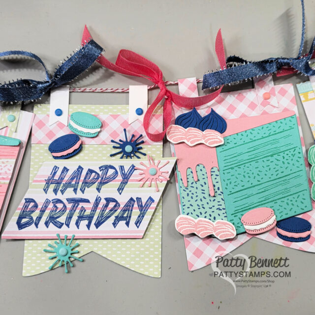 A handmade birthday banner with pastel flags featuring “HAPPY BIRTHDAY” in bold letters, colorful die-cut macarons, blue and pink ribbons, and decorative elements like swirls, dots, and faux icing. “PattyStamps.com” is visible at the bottom right. To read more, visit Patty Stamps (pattystamps.com)