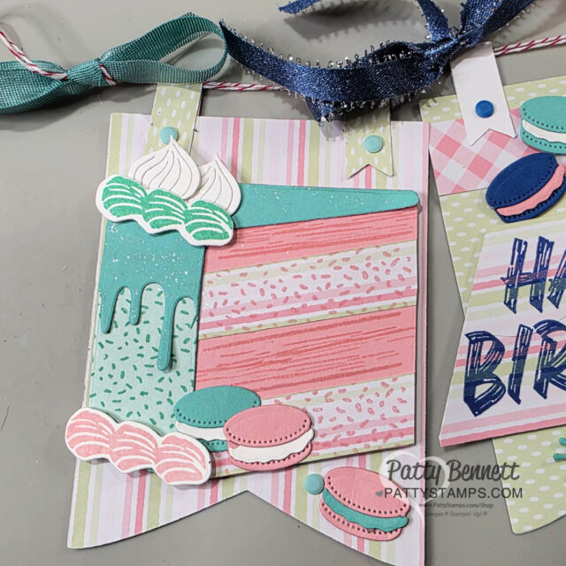 A decorative birthday banner features a pastel layer cake with mint green icing, sprinkles, and meringues, alongside pink and teal macarons. Behind it, the banner has pastel diagonal stripes and is topped with a mint green ribbon tied in a bow. To read more, visit Patty Stamps (pattystamps.com)