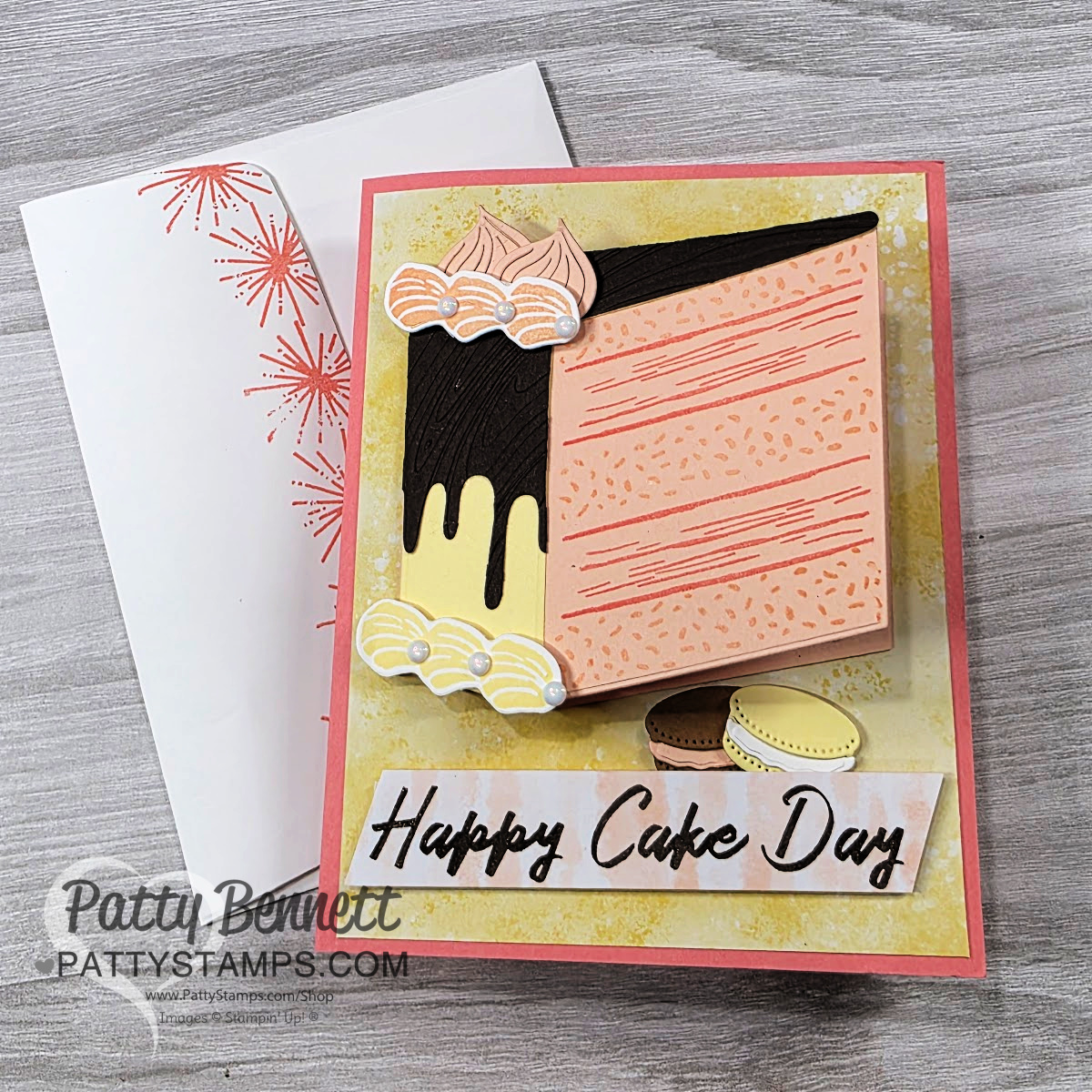 A handmade greeting card features a large, layered slice of cake with chocolate and yellow icing, whipped cream, and macarons. The card reads "Happy Cake Day" in black script and is set on a soft yellow and pink background, next to a white envelope with a red burst design. To read more, visit Patty Stamps (pattystamps.com)