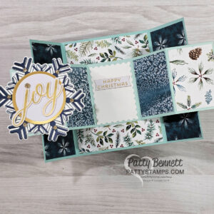 A handmade, three-panel Christmas card is displayed open on a wooden surface. It features blue and white botanical paper, a “JOY” snowflake embellishment on the left, and a “HAPPY CHRISTMAS” message in the center. The card is signed "Patty Bennett. To read more, visit Patty Stamps (pattystamps.com)