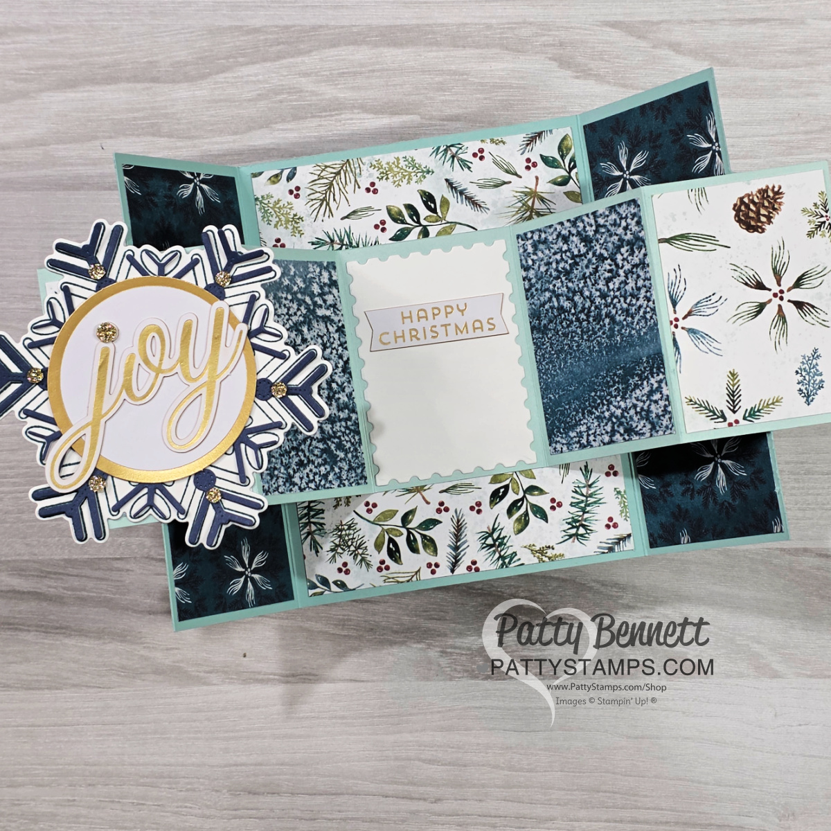 A handmade, three-panel Christmas card is displayed open on a wooden surface. It features blue and white botanical paper, a “JOY” snowflake embellishment on the left, and a “HAPPY CHRISTMAS” message in the center. The card is signed "Patty Bennett. To read more, visit Patty Stamps (pattystamps.com)