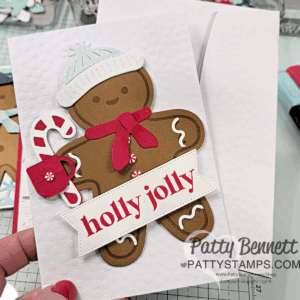 A hand holds a festive greeting card featuring a gingerbread figure with a white hat, red scarf, candy cane, and a "holly jolly" banner. The card has a textured white background. "Patty Bennett" and "PattyStamps.com" are watermarked at the bottom. To read more, visit Patty Stamps (pattystamps.com)