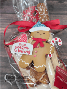 A gingerbread man card decorated with a red bow, white knit hat, and festive tag reading "'tis the season for baking" is attached to a cellophane-wrapped holiday baking gift with a candy cane, wooden spoon, and gingerbread baked good. To read more, visit Patty Stamps (pattystamps.com)