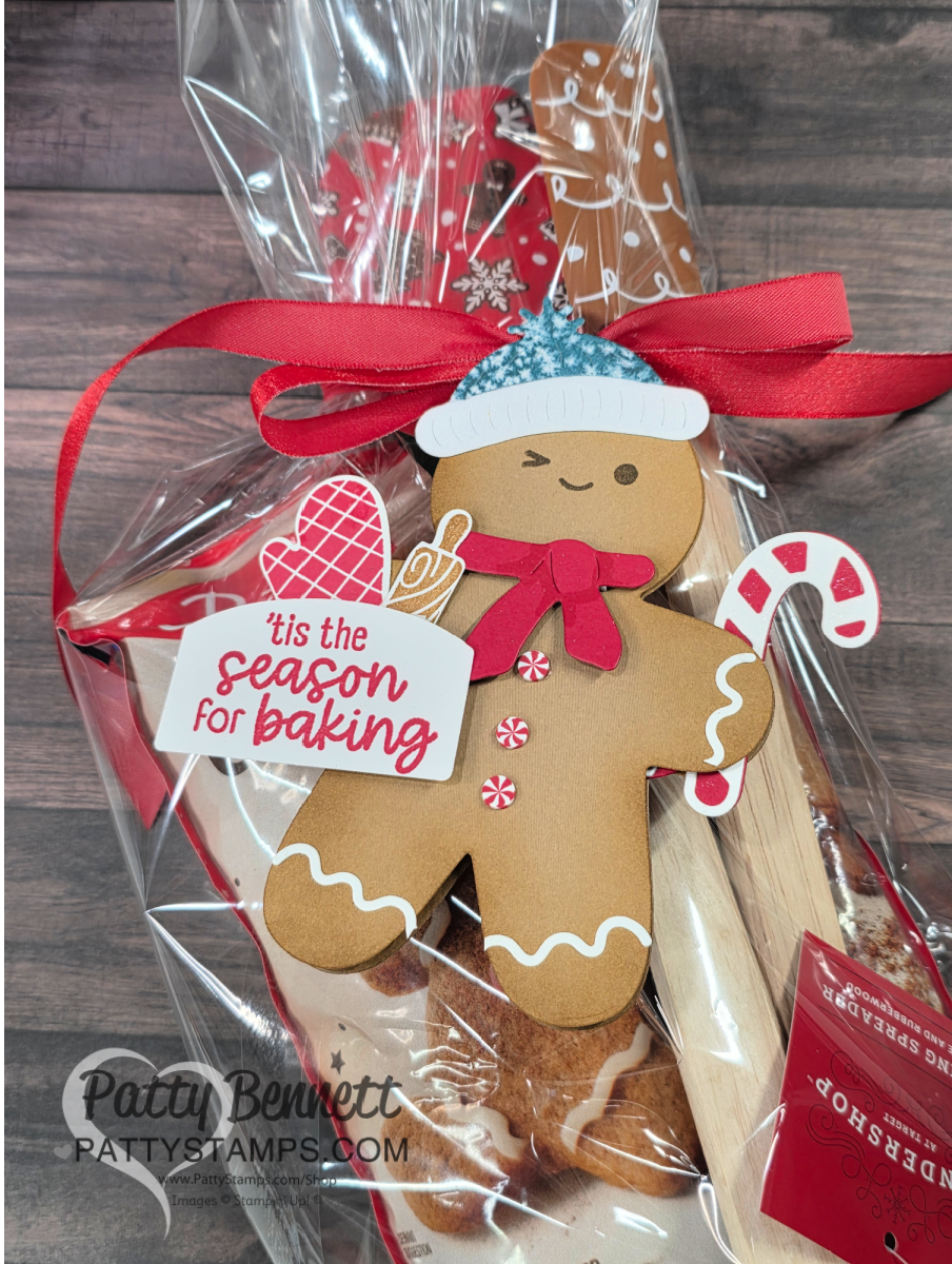 A gingerbread man card decorated with a red bow, white knit hat, and festive tag reading "'tis the season for baking" is attached to a cellophane-wrapped holiday baking gift with a candy cane, wooden spoon, and gingerbread baked good. To read more, visit Patty Stamps (pattystamps.com)