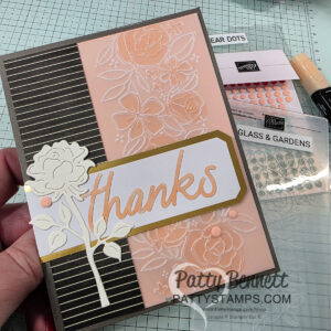 A handmade thank you card is held above craft supplies. The card has black-and-gold stripes, a peach floral embossed panel, a white die-cut flower, and "thanks" in peach script on a gold-bordered label. Stamps, embellishments, and tools are in the background. To read more, visit Patty Stamps (pattystamps.com)
