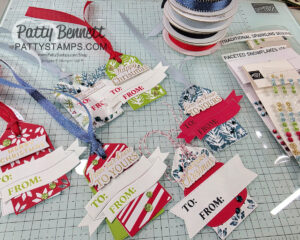A tabletop displays handmade Christmas gift tags with festive patterns, “TO” and “FROM” sections, and “Merry Christmas” or “Happy Christmas” banners. Ribbons, gem stickers, spools of ribbon, and crafting supplies surround the tags on a grid-patterned mat. To read more, visit Patty Stamps (pattystamps.com)