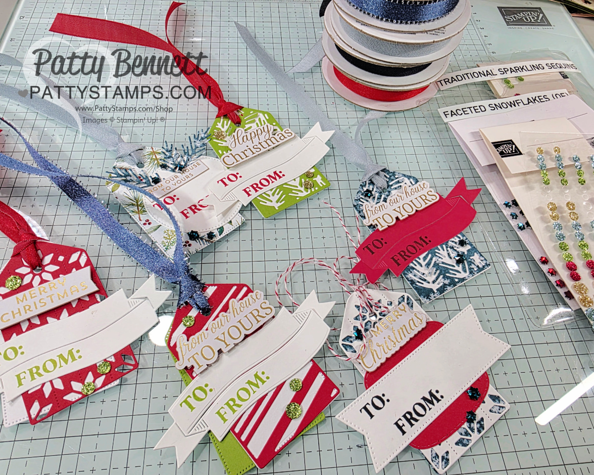 A tabletop displays handmade Christmas gift tags with festive patterns, “TO” and “FROM” sections, and “Merry Christmas” or “Happy Christmas” banners. Ribbons, gem stickers, spools of ribbon, and crafting supplies surround the tags on a grid-patterned mat. To read more, visit Patty Stamps (pattystamps.com)
