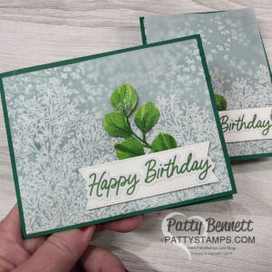 A handmade birthday card features embossed white foliage on a soft green background, green layered leaves in the center, and a white banner with "Happy Birthday" in green script. The card is held in hand; another similar card lies behind it. "PattyStamps" watermark appears. To read more, visit Patty Stamps (pattystamps.com)