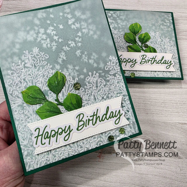 A hand holds a handmade birthday card featuring embossed white floral and leaf designs on a gray-green background. Green paper leaves and a “Happy Birthday” message in green script are layered on the front. Another matching card lies behind it. To read more, visit Patty Stamps (pattystamps.com)