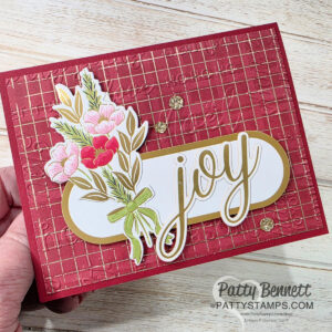 A handmade greeting card features a red embossed background with a gold grid pattern, a floral bouquet with pink and red flowers tied with a green bow, and a white oval label with the gold word “joy.” The card is held by a hand. To read more, visit Patty Stamps (pattystamps.com)