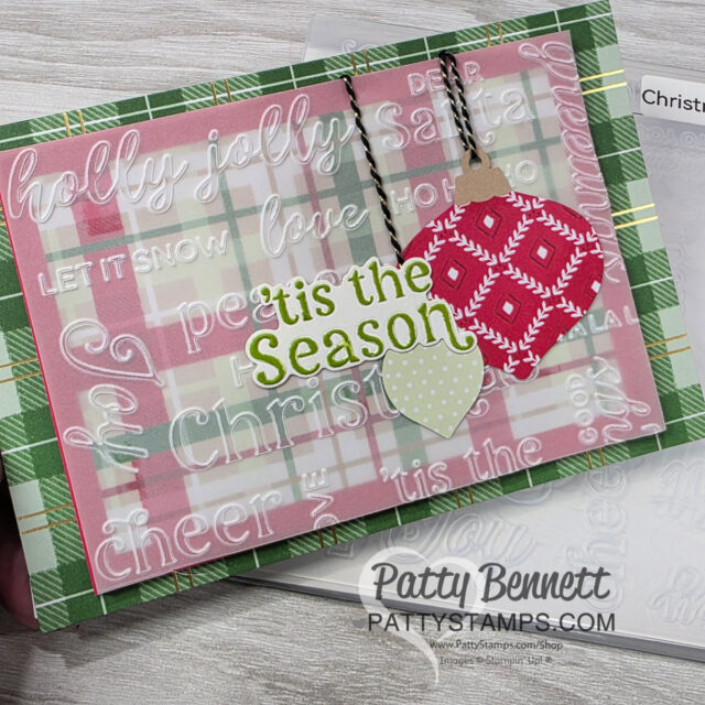 A handmade holiday card features layered plaid and pink backgrounds with white embossed festive words. Two paper ornaments, one red with a white knit pattern and one light green, hang by black cords. “‘Tis the Season” is stamped in green. “Patty Bennett” appears at the bottom. To read more, visit Patty Stamps (pattystamps.com)