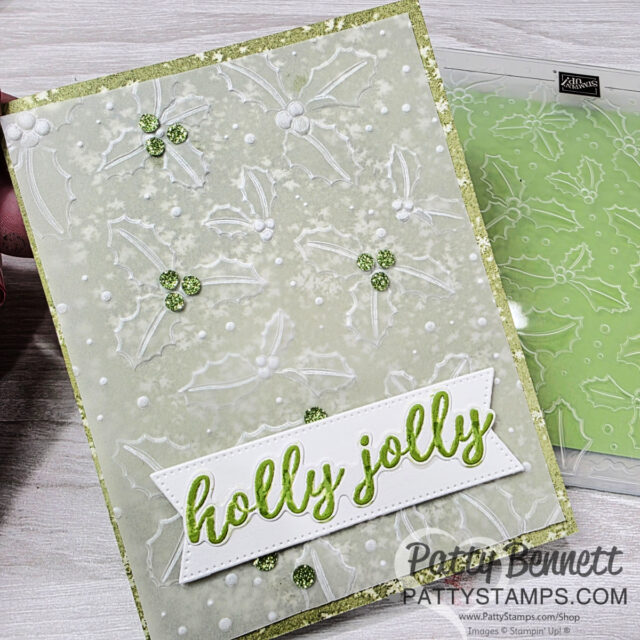 A handmade holiday card with embossed white holly leaves and berries on a pale green background. Glittery green accents highlight the berries. A white banner at the bottom reads “holly jolly” in green script. The PattyStamps watermark is in the lower right. To read more, visit Patty Stamps (pattystamps.com)