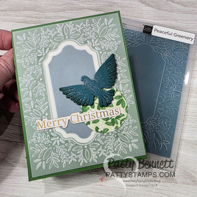 A handmade Christmas card features a raised, blue bird and “Merry Christmas” text on a decorative tag. The pale green card base has an embossed leafy border and is paired with a “Peaceful Greenery” embossing folder in the background. A hand holds the card. To read more, visit Patty Stamps (pattystamps.com)