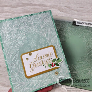 A handmade card with a frosty white leaf pattern on a light green background. A gold-edged label reads "Season's Greetings" in gold, decorated with holly leaves and berries. Three gold glitter dots accent the card. A matching embossing folder is shown beside it. To read more, visit Patty Stamps (pattystamps.com)