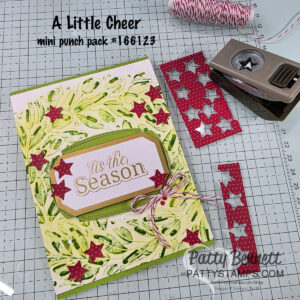 A handmade holiday card with green leafy textured background, gold “’tis the Season” label, and red star accents. Nearby are red star-patterned paper strips, a star-shaped paper punch, red-and-white twine, and the text: “A Little Cheer mini punch pack #166123.”. To read more, visit Patty Stamps (pattystamps.com)