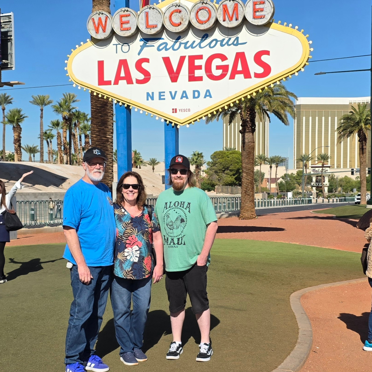 Three people stand smiling in front of the iconic "Welcome to Fabulous Las Vegas Nevada" sign. Palm trees and buildings are in the background on a sunny day. The group, two men and one woman, are casually dressed and stand on a green patch of artificial grass. To read more, visit Patty Stamps (pattystamps.com)
