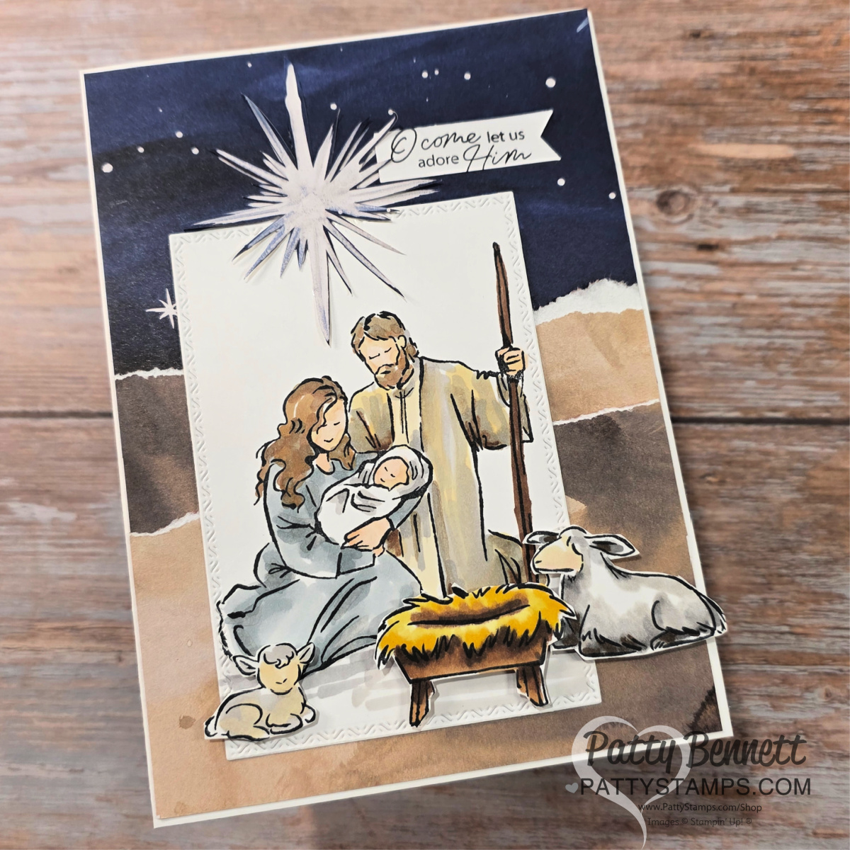 A handmade Christmas card depicts the Nativity scene with Mary, Joseph, and baby Jesus in a manger, surrounded by two lambs. A bright star shines above with the words “O come let us adore Him.” The background shows a night sky and rocky landscape. To read more, visit Patty Stamps (pattystamps.com)