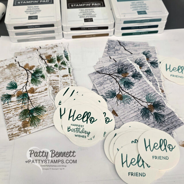 Multiple handmade card components are shown: rectangular pieces with pine branch designs, circular tags reading “Hello FRIEND” and “Hello HAPPIEST BIRTHDAY WISHES.” Several stacked ink pads and the watermark “Patty Bennett PattyStamps.com” are visible in the foreground. To read more, visit Patty Stamps (pattystamps.com)