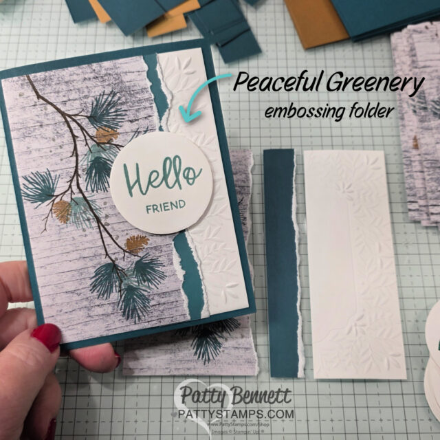 A hand holds a handmade card with pine branch art, a "Hello Friend" circle, torn teal and white embossed strips. Text reads "Peaceful Greenery embossing folder." Art supplies and card pieces are scattered on a grid table. "Patty Bennett" watermark is at the bottom. To read more, visit Patty Stamps (pattystamps.com)