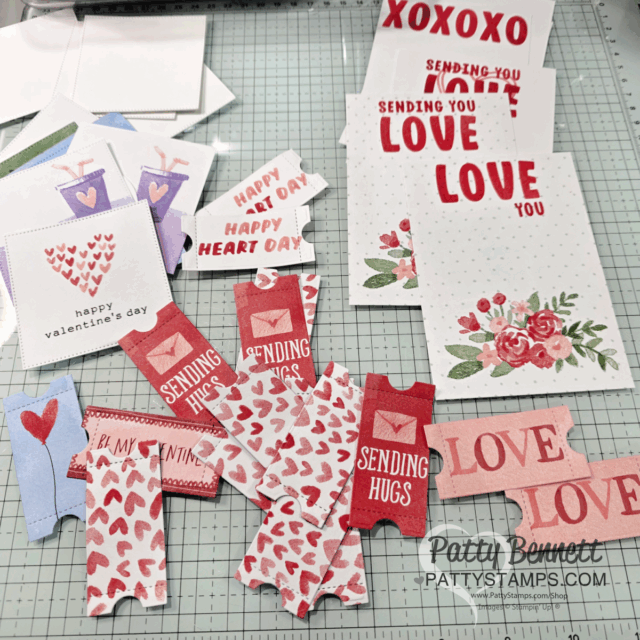 A craft table displays handmade Valentine cards, fun fold cards, and paper cutouts with hearts, “LOVE,” “Sending Hugs,” and “Happy Valentine’s Day.” Designs feature red, pink, and floral patterns. Some cards are assembled; others are in progress. To read more, visit Patty Stamps (pattystamps.com)