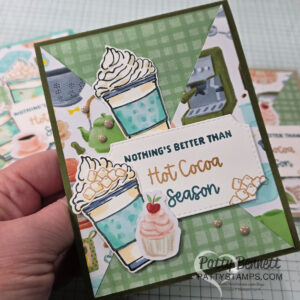 A hand holds a handmade greeting card decorated with drawings of hot cocoa cups topped with whipped cream and marshmallows, a cupcake, and festive accents. The card reads, "NOTHING'S BETTER THAN Hot Cocoa Season" in green, brown, and orange text. To read more, visit Patty Stamps (pattystamps.com)