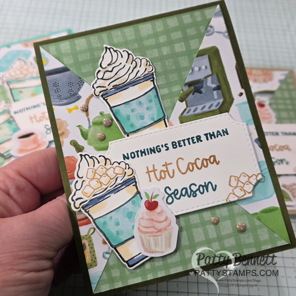 A hand holds a handmade greeting card decorated with drawings of hot cocoa cups topped with whipped cream and marshmallows, a cupcake, and festive accents. The card reads, "NOTHING'S BETTER THAN Hot Cocoa Season" in green, brown, and orange text. To read more, visit Patty Stamps (pattystamps.com)