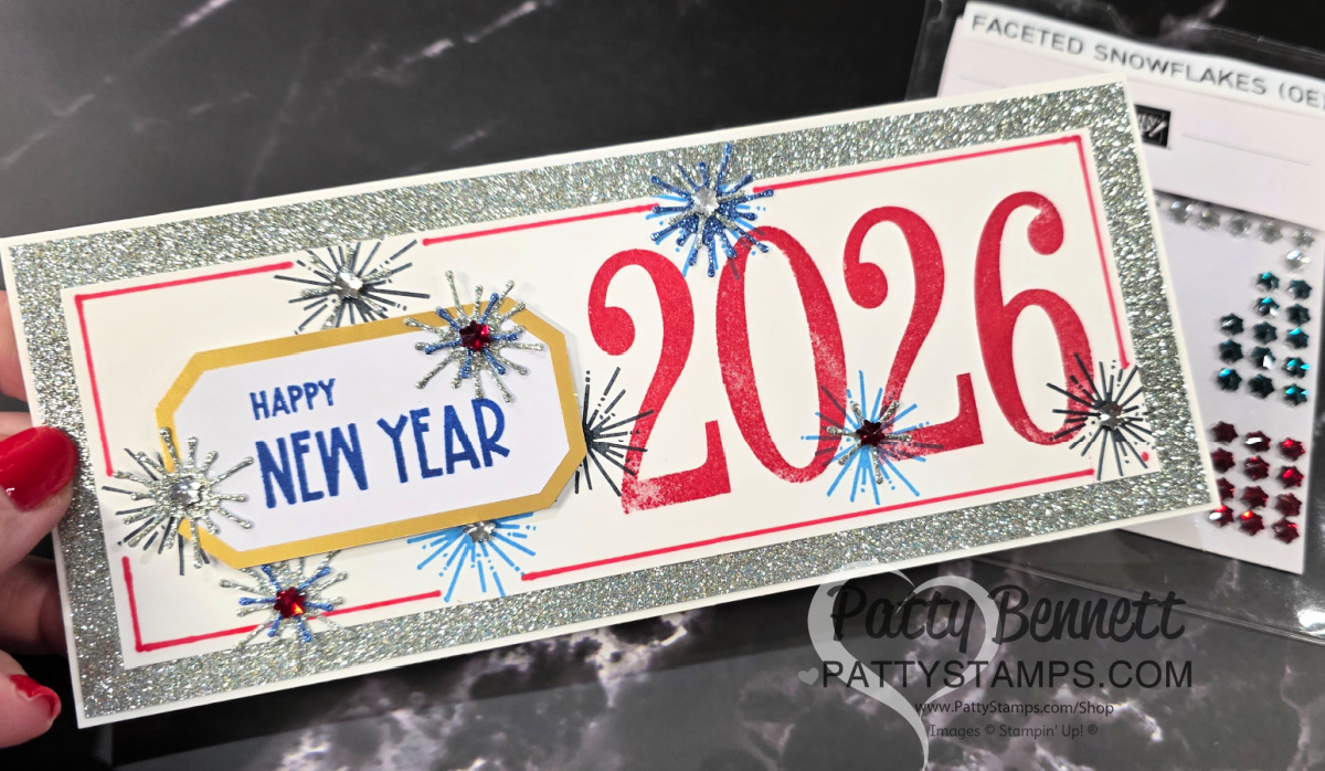 A handmade New Year card with large red "2026" numbers, blue starbursts, silver glitter border, and a yellow label reading "HAPPY NEW YEAR" in blue text. The card is held by a hand with red nails. Faceted snowflake gems are visible in the background. To read more, visit Patty Stamps (pattystamps.com)