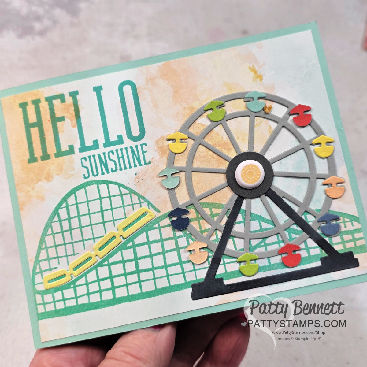 A hand holds a cheerful greeting card featuring a colorful Ferris wheel, a roller coaster, and the text “HELLO SUNSHINE” in bold yellow and teal letters. The card has a soft, pastel background and is signed “Patty Bennett PattyStamps.com.”. To read more, visit Patty Stamps (pattystamps.com)