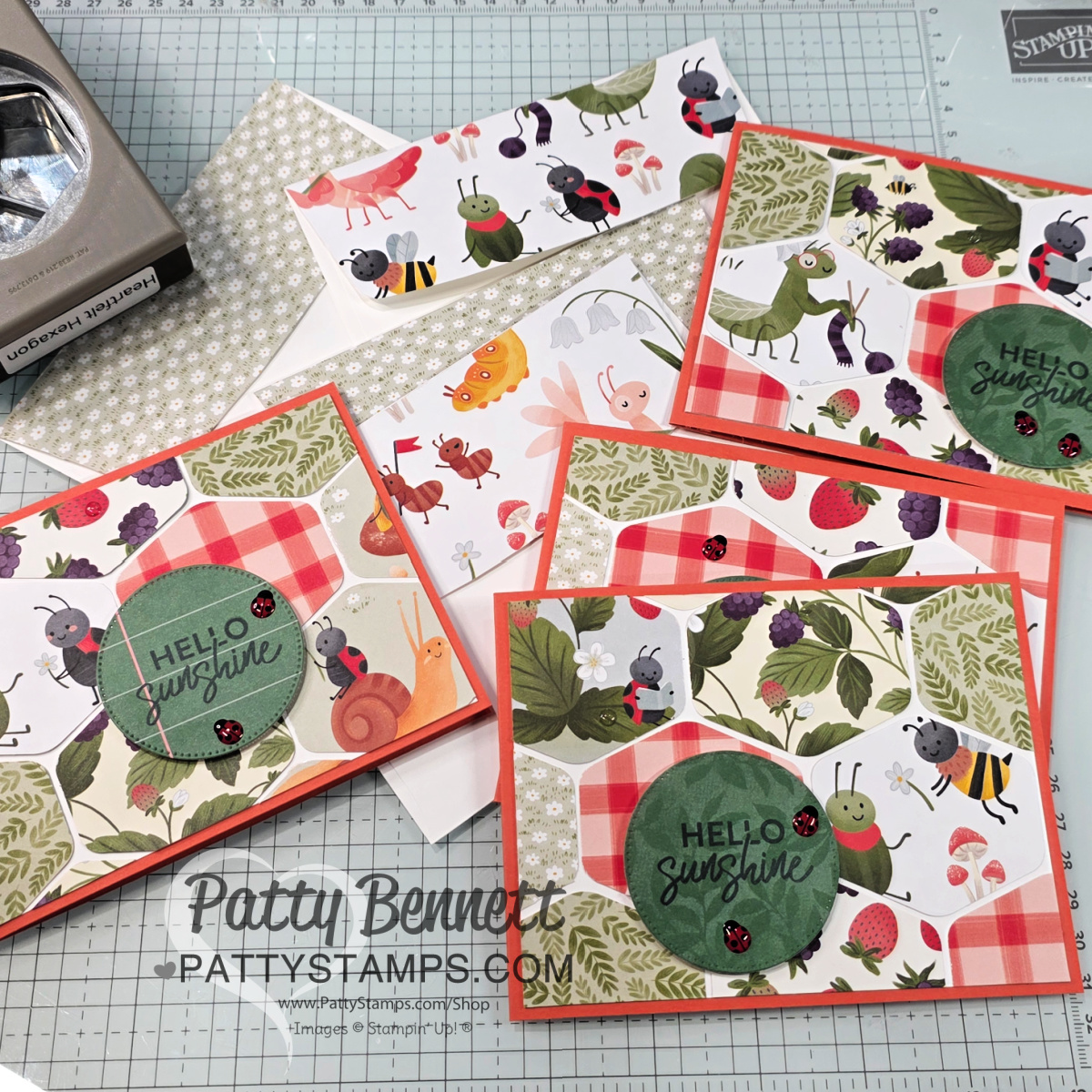 A set of handmade greeting cards with animal and fruit-themed patterned paper, red borders, and round green labels reading "HELLO sunshine." The cards are arranged on a craft mat with tools and materials scattered around, including an envelope and a paper punch. To read more, visit Patty Stamps (pattystamps.com)